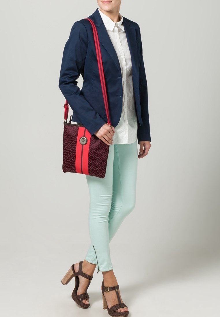 Červeno-modrá taška přes rameno s texturovaným vzorem, vybavená stříbrným logem a výraznými červenými pruhy. Modelka má na sobě tmavě modrý blejzr a mintové kalhoty.