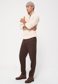 Kremowy, teksturowany sweter na zamek; brązowe, dopasowane spodnie; brązowe sznurowane buty; jasna dzianinowa czapka; model stoi z rękami skrzyżowanymi na tle białym.