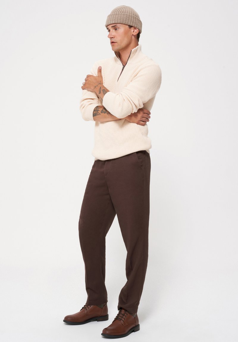 Kremowy, teksturowany sweter na zamek; brązowe, dopasowane spodnie; brązowe sznurowane buty; jasna dzianinowa czapka; model stoi z rękami skrzyżowanymi na tle białym.