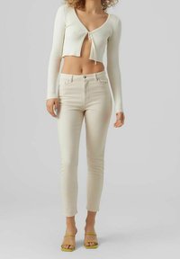 Krémový lehký ribbed krácený cardigan s výstřihem do V a řetízkovým detailem, spárovaný se světle béžovými skinny džíny s vysokým pasem a zelenými sandály na řemínky.
