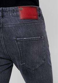 Pantalones vaqueros de denim lavado oscuro con una apariencia texturizada y desteñida; cuentan con una parche de cuero rojo en la cintura con el nombre de la marca en relieve.
