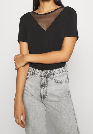 Kobieta ubrana w czarny krótki top z przezroczystą, siateczkową wstawką w kształcie litery V, zestawiony z jasnoszarymi, wysokim stanem jeansami.