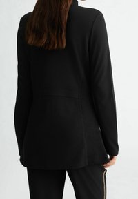 Czarna kurtka z długim rękawem o dopasowanym kroju, gładkiej fakturze i zaokrąglonym dolnym brzegu, z wysokim kołnierzem i bez widocznych zapięć.