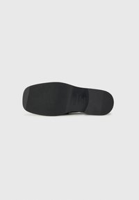 Schwarze Schuhsohle mit einem strukturierten, gerippten Muster. Das Material erscheint gummiartig, flach mit leichter Krümmung an der Zehenpartie. Kleines Branding-Detail sichtbar.