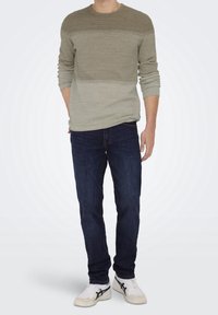 Stickad tröja i olivgrön och ljusgrön med en texturerad yta, tillsammans med mörkblå jeans och vita sneakers med svarta accenter.