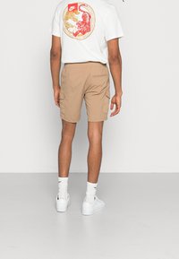 Bílý tričko s grafickým motivem červeného a zlatého tygra na zádech, béžové cargo šortky s bočními kapsami a bílé tenisky s žebrovanými ponožkami.