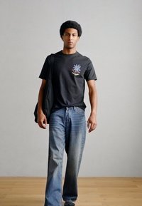 Μαύρο βαμβακερό t-shirt με γραφικό σχέδιο στο στήθος, σε συνδυασμό με μπλε τζιν παντελόνι. Το μοντέλο κρατάει μαύρη τσάντα ώμου.