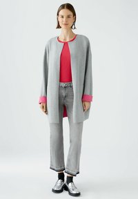 Helle graue Strickjacke mit leuchtend pinken Bündchen, getragen über einem roten Oberteil und grauen Jeans. Sie hat seitliche Taschen und eine entspannte Passform. Silberne Loafers runden das Outfit ab.