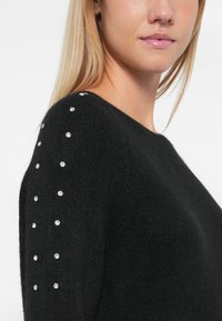 Sweater preto de malha com decote redondo e tecido texturizado, adornado com tachas prateadas ao longo dos ombros.