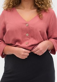 Blouse rose texturée avec un décolleté en V profond, des manches trois-quarts et quatre boutons ronds sur le devant. Associée à une jupe noire.