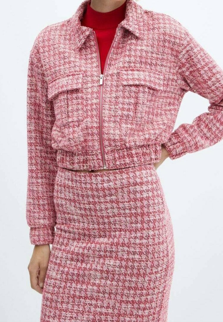 Femme portant une veste zippée à carreaux rose et blanc avec poches à rabat, assortie à une jupe, et un haut à col roulé rouge.