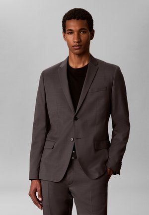 Homme portant une veste et un pantalon de costume gris foncé ajustés avec une chemise noire, debout devant un fond gris clair uni.