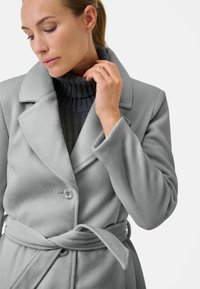 Ljust grå fleecejacka med notch-krage, enkel knappstängning och knytskärp. Har en texturerad ribbad polokrage under.