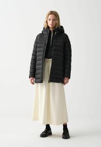 Černá péřová bunda s kapucí, prošívaný design, přední zip, nošená přes béžovou sukni. Základní černé boty dotvářejí outfit.