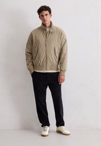 Beżowa kurtka bomberka z wysokim kołnierzem, zamkiem błyskawicznym z przodu i dwoma bocznymi kieszeniami, w połączeniu z granatowymi sztruksowymi spodniami i białymi trampkami. Gładka tekstura.