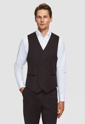 Mężczyzna w białej koszuli, czarnej dwurzędowej kamizelce i czarnych eleganckich spodniach, stojący na jednolitym jasnym tle.
