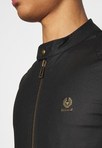 Czarna kurtka z wysokim kołnierzem, złotym zamkiem błyskawicznym i zapięciem na zatrzask. Posiada emblem markowy na piersi oraz gładką tkaninę.