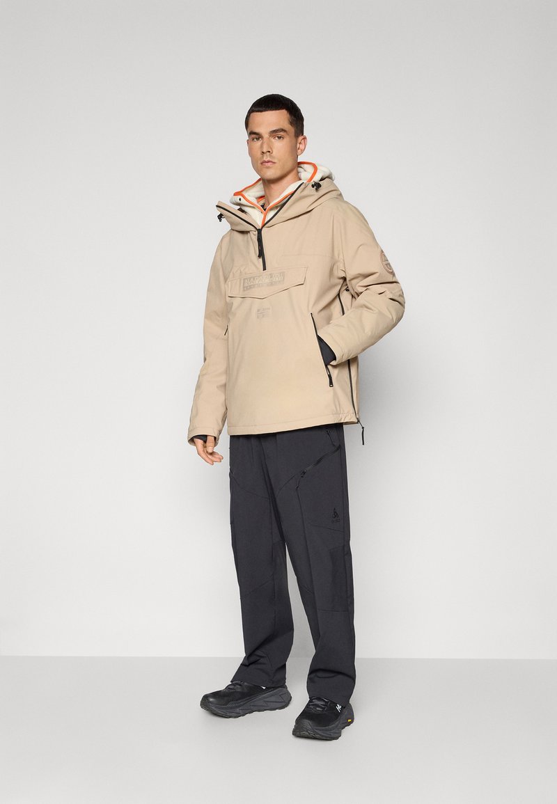 Veste à capuche beige avec fermeture éclair et cordon de serrage, équipée d'une poche avant. Associée à un pantalon cargo noir et des baskets noires.