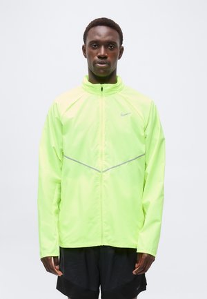 Mężczyzna w neonowo-żółtej sportowej kurtce na pełny zamek z odblaskowymi elementami i czarnych szortach, stojący na jednolitym białym tle.