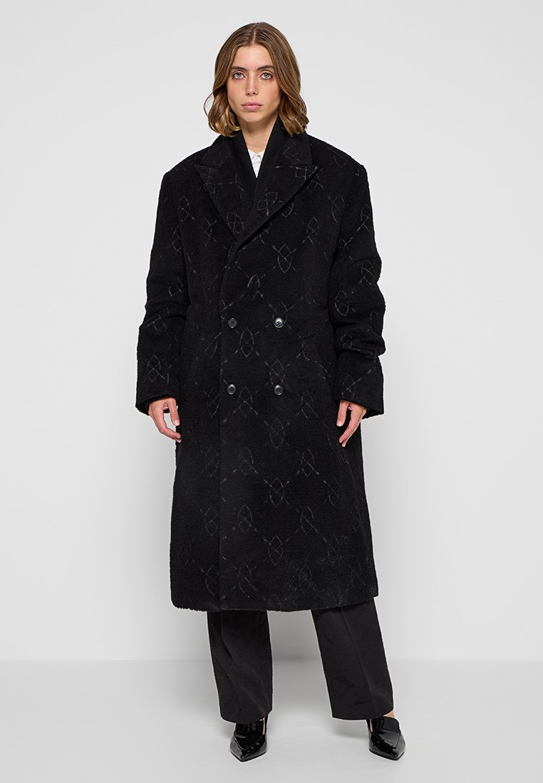 Manteau en mélange de laine noire avec un motif texturé, devant double boutonnage, grands revers et poches latérales. Le modèle porte un pantalon et des chaussures noirs.