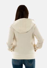 Felpa crema con cappuccio regolabile, maniche lunghe e una texture morbida, abbinata a jeans blu con dettagli di cucitura sulle tasche posteriori.