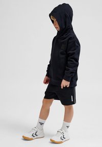 Svart huvtröja med framficka och logotyp, tillsammans med svarta shorts med sidostreck, vita sportiga sneakers och knähöga strumpor.