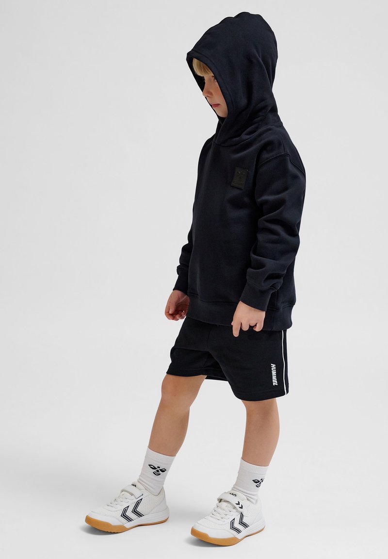 Svart huvtröja med framficka och logotyp, tillsammans med svarta shorts med sidostreck, vita sportiga sneakers och knähöga strumpor.