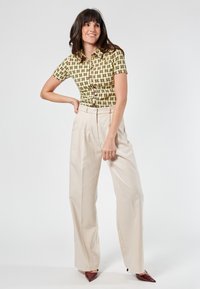 Top verde a maniche corte con un motivo geometrico, abbinato a pantaloni beige a vita alta e scarpe borgogna a punta, in piedi su uno sfondo chiaro.