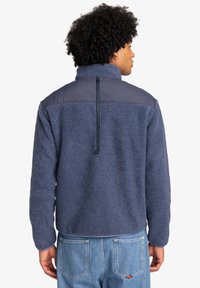 Navy blue fleece jacket with a high collar, textured fabric, and a smooth upper panel. Features a zip at the back and elastic cuffs.