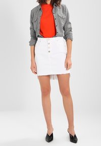 Gonna di jeans bianca con orlo sfilacciato e bottoni frontali, abbinata a una maglietta rossa e una camicia a quadri nera. Ballerine nere a punta.