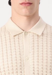 Polo din tricot bej cu un model texturat, guler moale și nasturi crem, cu un design slim fit și material ușor.