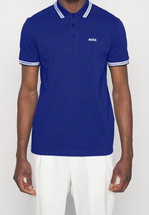 Mann in einem königsblauen Poloshirt mit einem weißen und blauen gestreiften Kragen und Ärmeln, der entspannt mit den Armen an den Seiten steht.