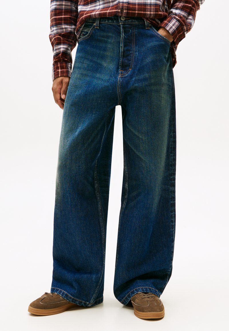 Jeans a gamba larga in denim blu scuro, con vita alta, taglio dritto e cuciture a contrasto. Abbinati a scarpe marroni.