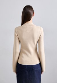 Pulover tricotat bej cu mâneci lungi, design ajustat și un mic detaliu de accent pe spate. Purtați cu o fustă din denim închis.