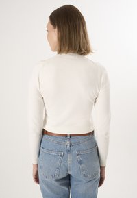 Kremfarvet, tætsiddende sweater med lange ærmer, båret sammen med blå højttaljede jeans. Bagsiden har et enkelt design med en let tekstur.
