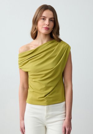 Groene top met een schouderontwerp, gemaakt van gladde stof; beschikt over gestructureerde plooien en een aansluitende pasvorm. Gedirigeerd met witte broek.