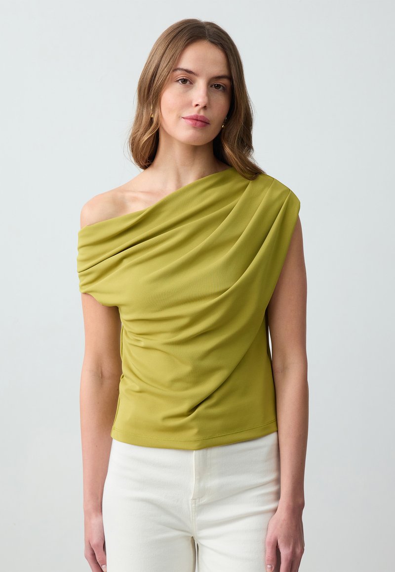 Haut drapé vert avec un design à une épaule, fabriqué en tissu lisse ; présente des plis texturés et une coupe ajustée. Porté avec un pantalon blanc.