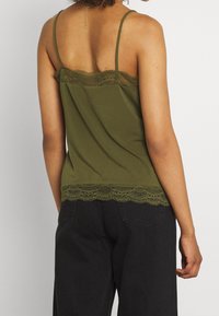 Femme portant un débardeur olive avec des bordures en dentelle et un pantalon noir taille haute, photographiée de dos sur fond uni.