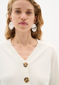 Krämbakad cardigan med ribbad V-ringning, prydd med tre stora, runda knappar i sköldpaddsmönster; modellen bär glänsande, överdimensionerade örhängen.