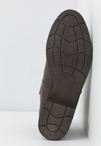 Semelle d'une chaussure marron avec une bande de roulement en caoutchouc noir à motifs, taille 36, positionnée verticalement contre un mur d'angle blanc.