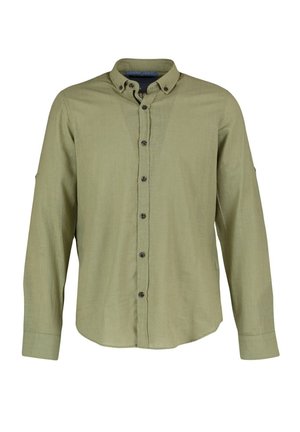 Man draagt een lichtgroen overhemd met lange mouwen en donkere broek, staand met één hand in de zak tegen een witte achtergrond.