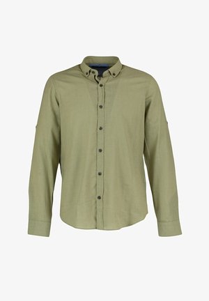 Man draagt een lichtgroen overhemd met lange mouwen en donkere broek, staand met één hand in de zak tegen een witte achtergrond.