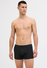 Young man with short hair wearing black boxer briefs standing against a plain light background, looking slightly to the side.