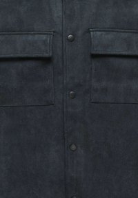 Giacca in suede nera con due tasche sul petto, chiusura con bottoni e texture liscia. Il davanti è rifinito con bottoni a scatto neri.