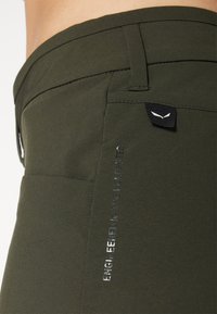 Pantalones cargo de tela verde oscuro con un diseño elegante, que cuentan con un bolsillo lateral prominente y texto reflectante que dice "INGENIERÍA EN LOS DOLOMITAS."