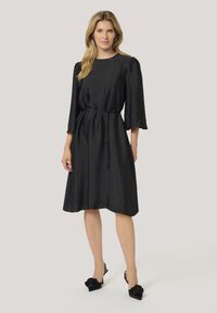 Robe noire à mi-mollet avec une finition texturée, manches trois-quarts, taille cintrée et décolleté rond. Associée à des talons à bride arrière noirs.