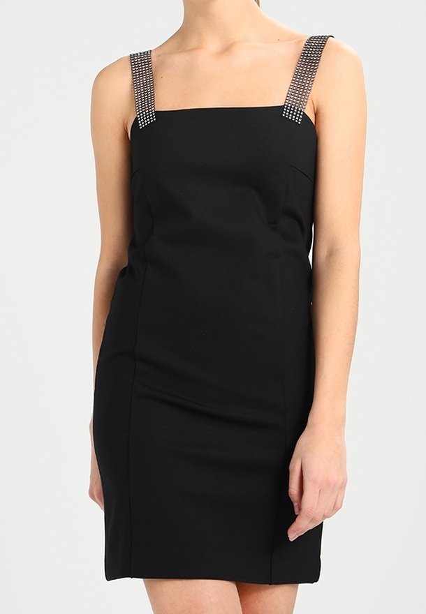 Femme portant une robe noire sans manches avec un décolleté carré et des bretelles ornées sur un fond clair uni.