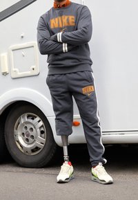 Sudadera y pantalones jogger grises de Nike con logo naranja, puños a rayas y una pierna prostética visible. Llevado con zapatillas Nike, de pie al lado de un vehículo.