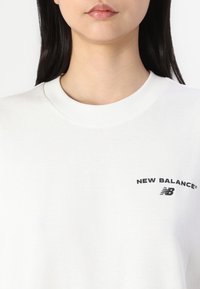 White cotton t-shirt featuring a crew neckline and a small black "New Balance" logo on the left chest area. Smooth texture, short sleeves.