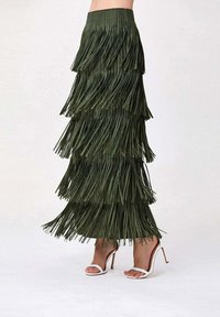 Jupe maxi verte à franges avec un design superposé, présentant une finition texturée et une taille élastique. Assortie à des sandales blanches à talons hauts.
